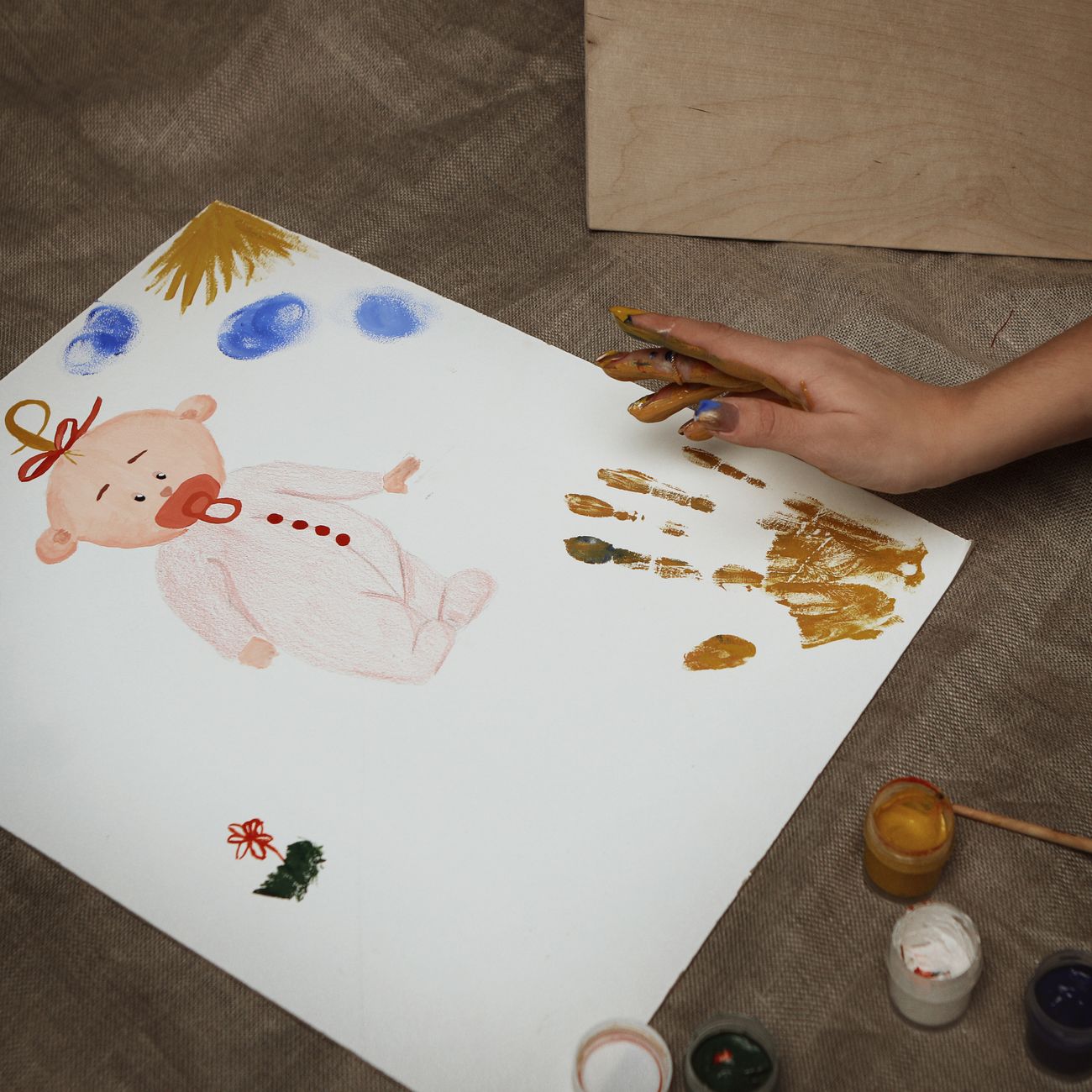 A child's golden handprints and a painted baby drawing on paper, with small jars of paint beside it