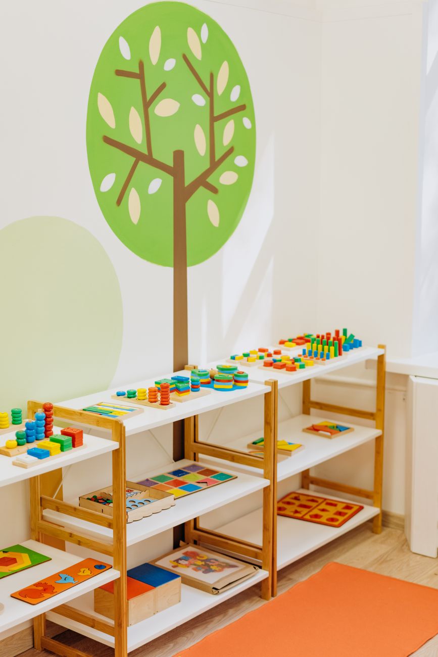 A low wooden classroom shelf holding colourful wooden puzzles and counting toys beneath a painted tree on the wall