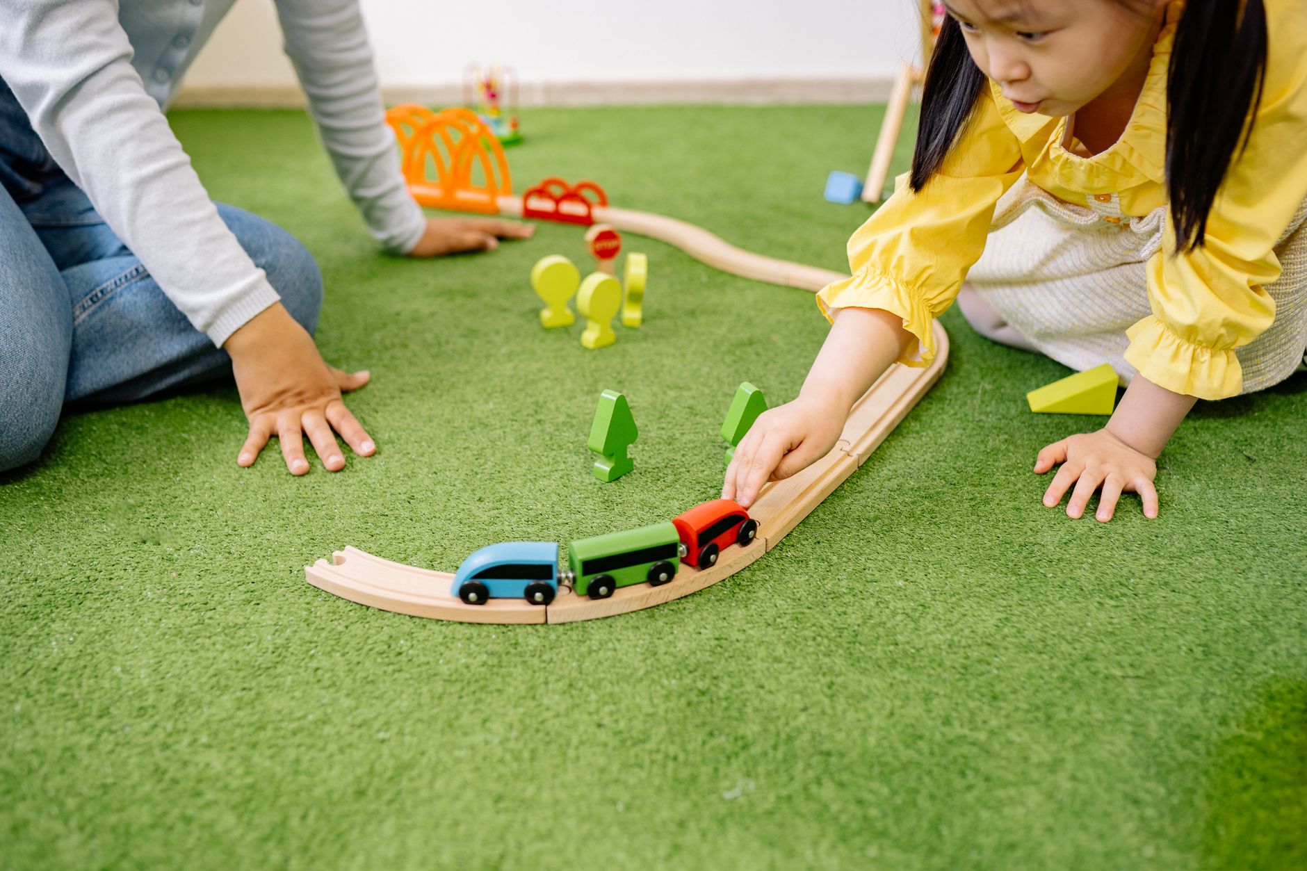 A child laying wooden train track on a green play mat while an adult's hand helps