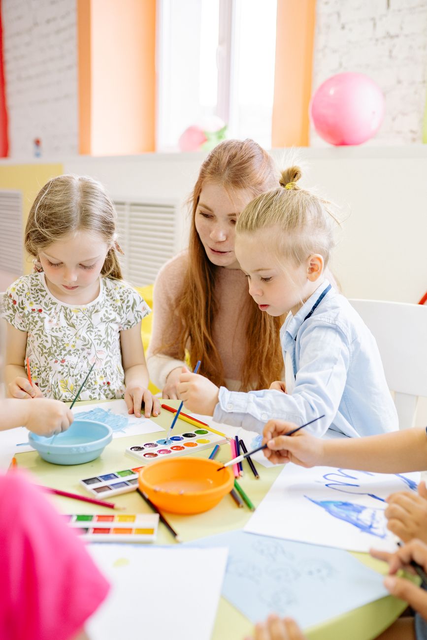 A teacher and two young children painting with watercolours at a low table in a sunny room
