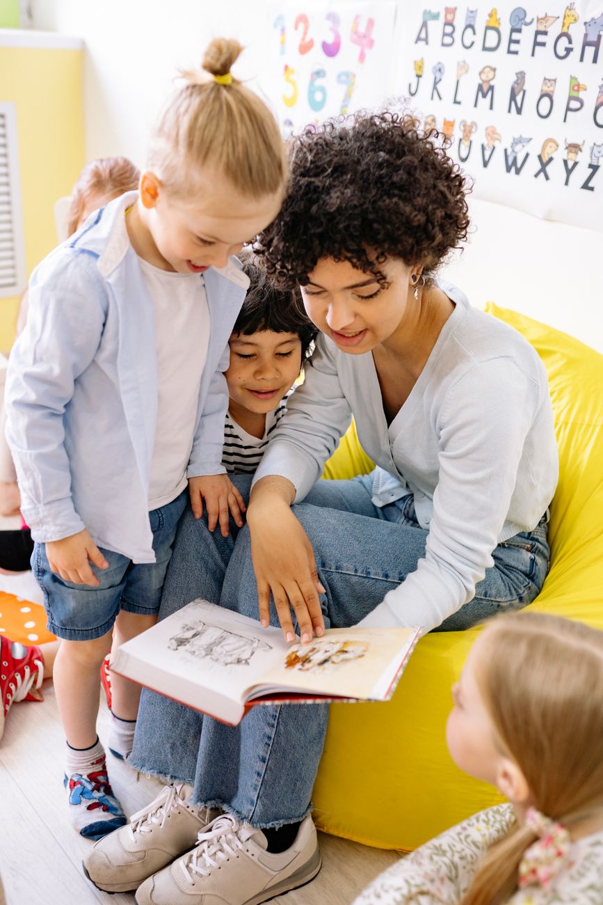 A teacher reading a picture book with children gathered around her on a yellow beanbag