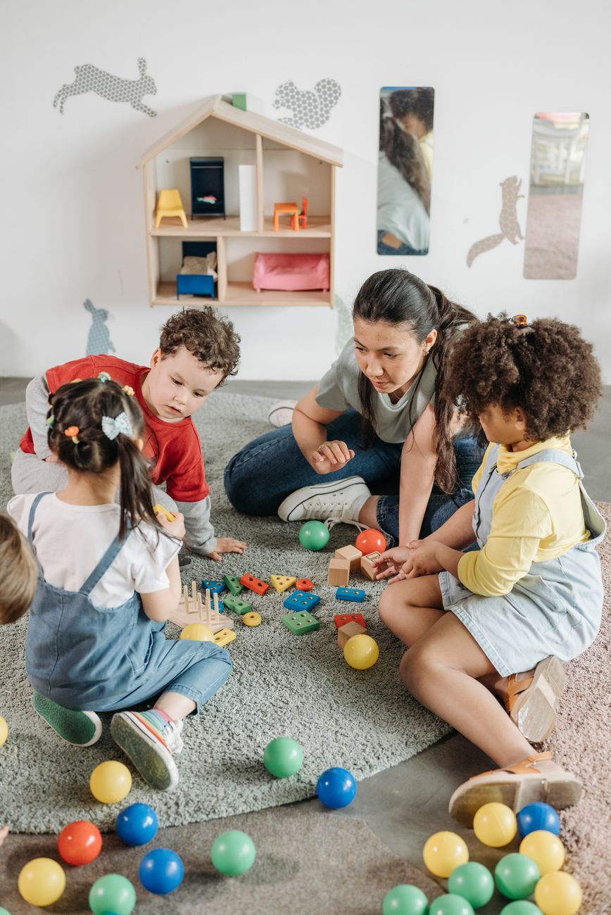 A teacher sitting on a soft rug with four preschool children playing with colourful balls and a small wooden dollhouse