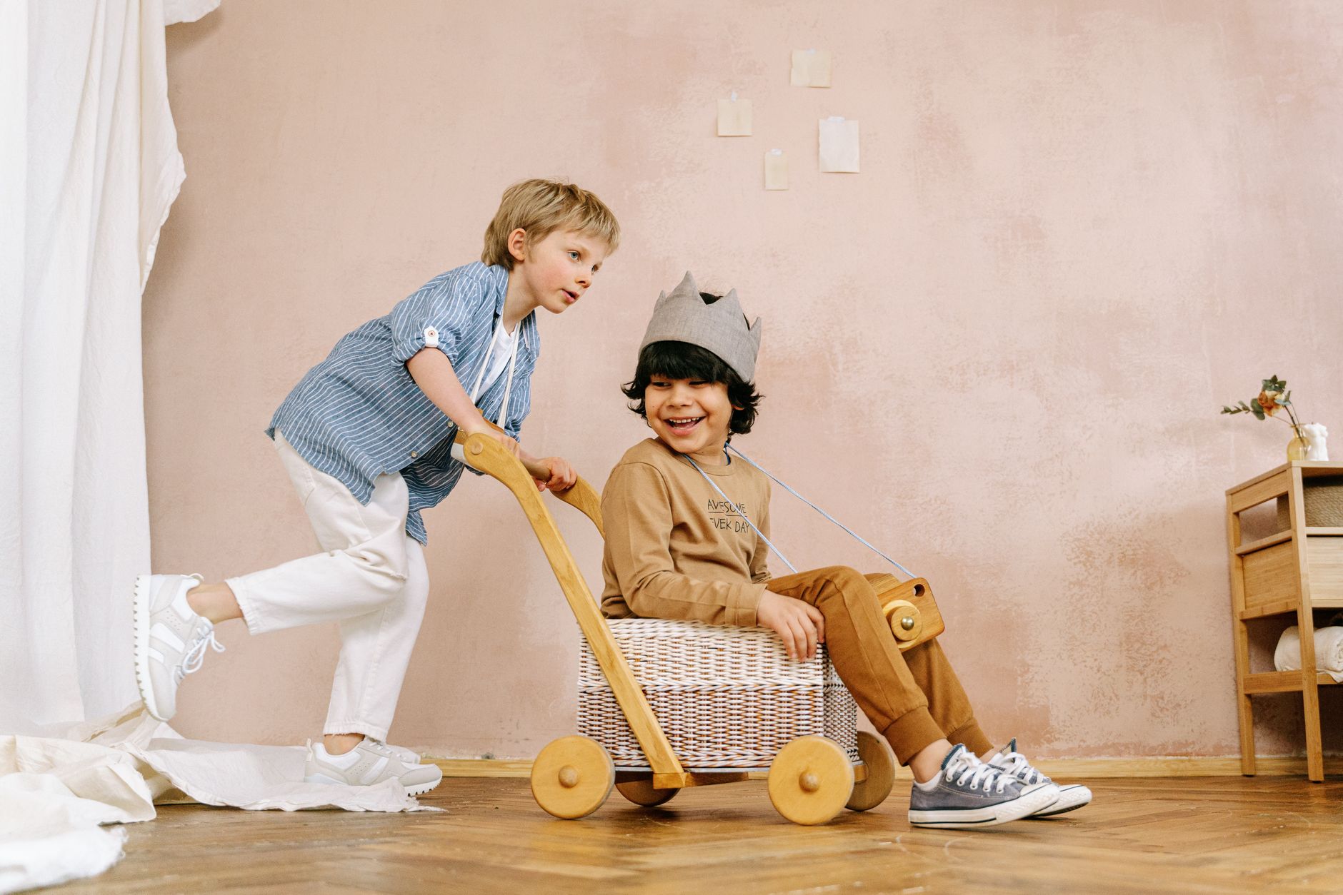 Two children playing a make-believe game with a wooden walker, one wearing a paper crown