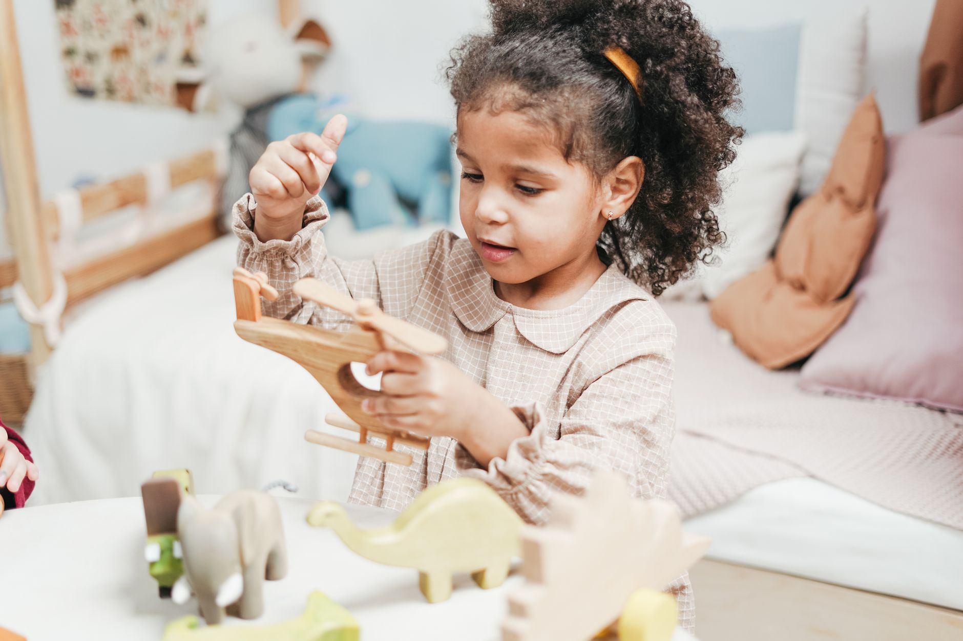 A curly-haired girl holding a wooden toy animal, with more wooden animals on the table in front of her