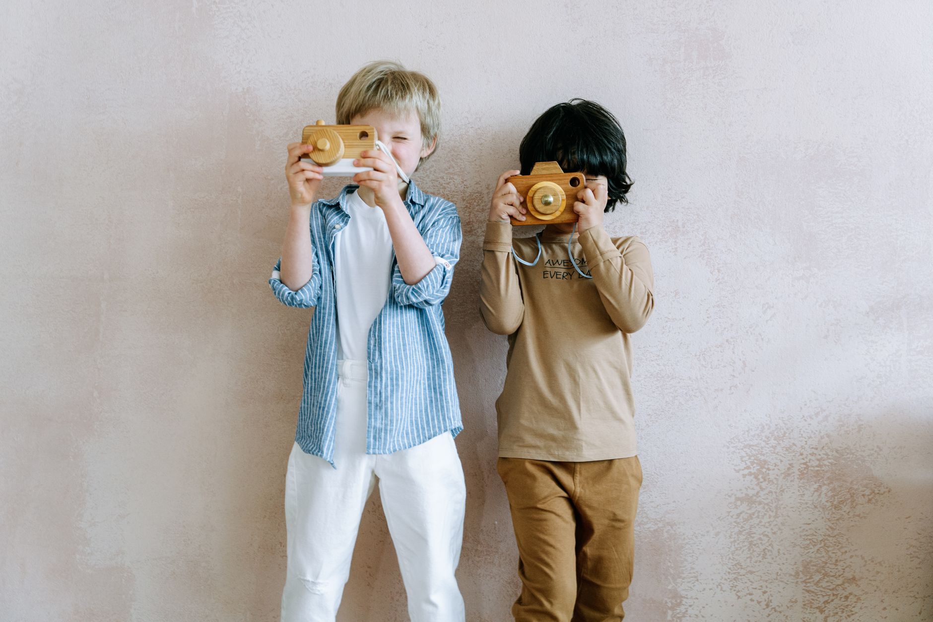 Two young boys lifting wooden toy cameras to their eyes against a soft pink wall