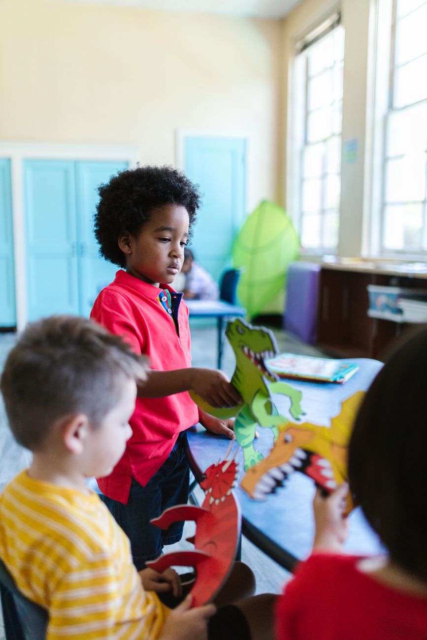 Three young children at a table in a bright classroom holding paper dinosaur cut-outs they made